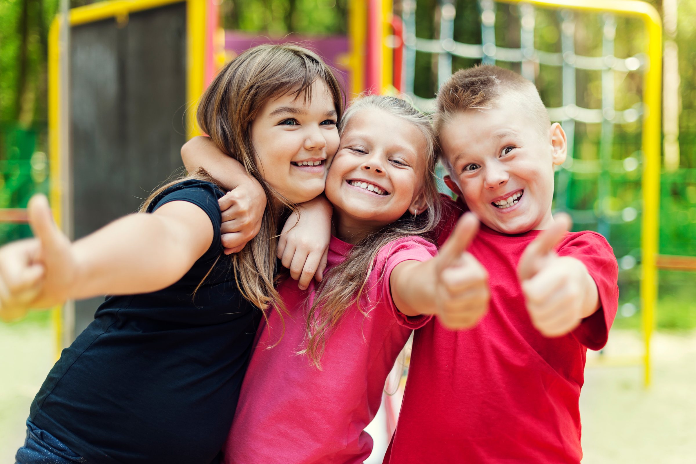 Drei fröhliche Kinder auf einem Spielplatz zeigen Daumen hoch